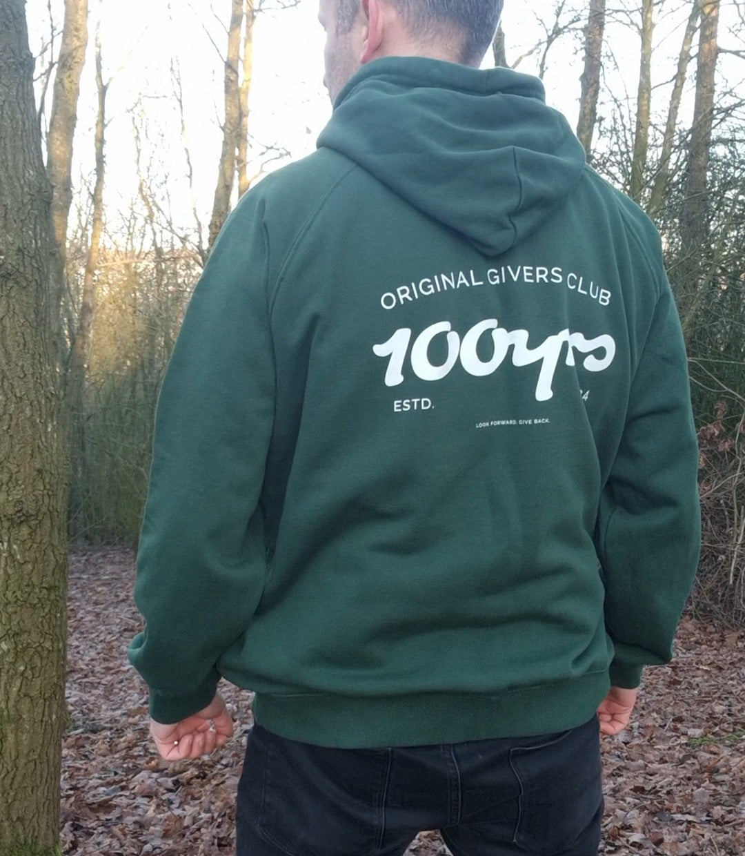 A man stood in a wooded area and wearing a green 100yrs organic cotton raglan hoodie.