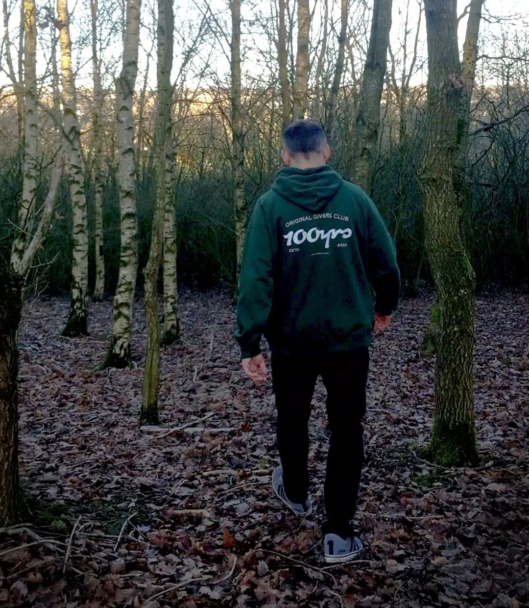 A man walking through a wooded area and wearing a green 100yrs organic cotton raglan hoodie.