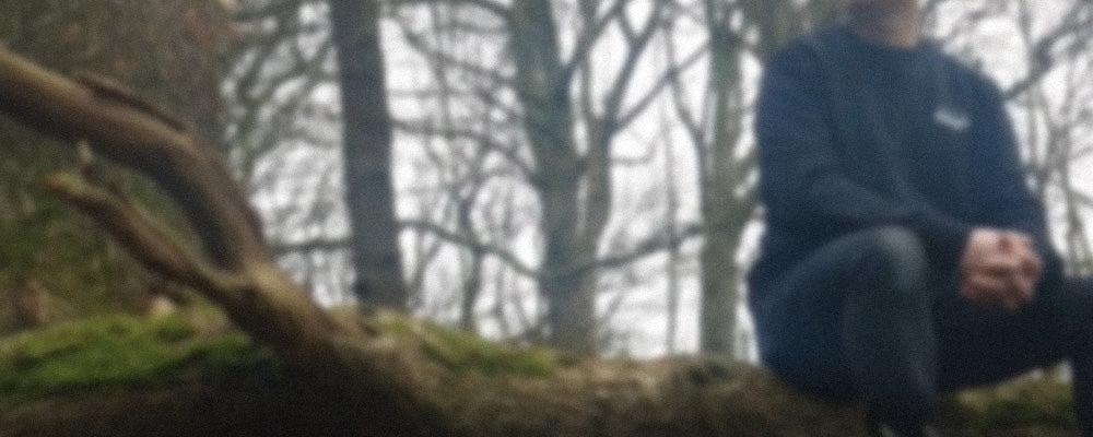 A man sitting in a wooded area wearing a black 100% organic cotton sweatshirt.
