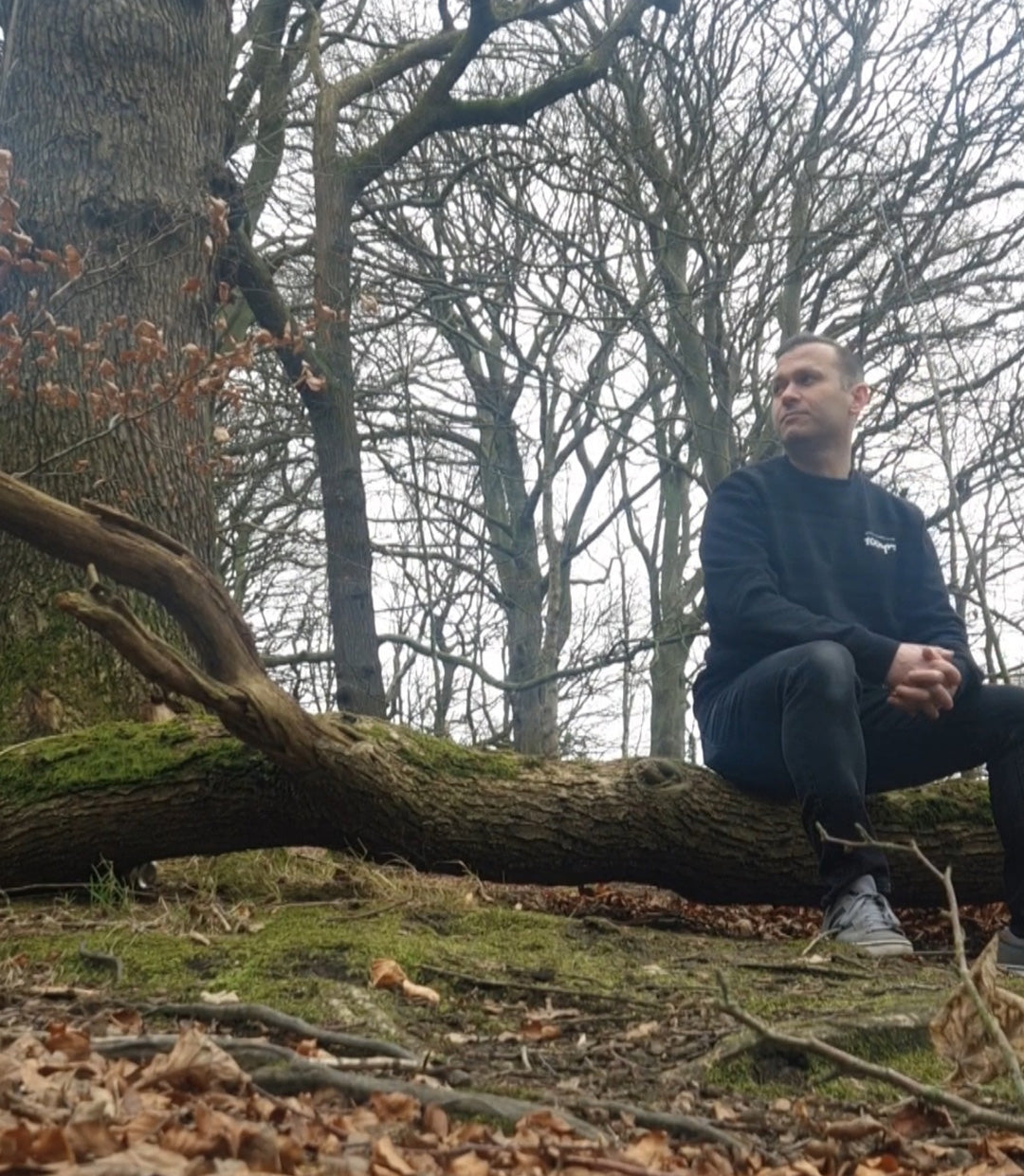 A man sitting on a fallen tree and wearing a black organic cotton sweatshirt.
